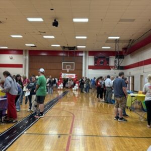 A school gym filled with people browsing tables set up along the sides for a community fair, balloons in the far end center.