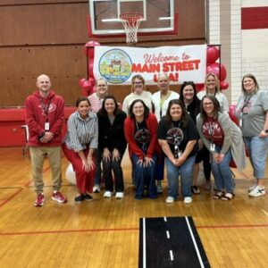 Group of adults posing for a photo in a school gym in front of a 'Welcome to Main Street' banner with red balloons, smiling at the camera.