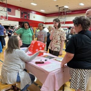 Woman seated at a table with a pink tablecloth in a gym, rolling a large red dice for a group of kids gathered around.
