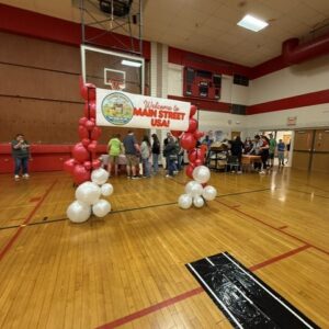 Arch of pink and white balloons frames a banner that says 'Welcome to Main Street USA!' in a school gym with a basketball hoop and people milling about in the background.