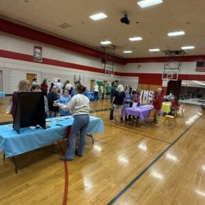 Community fair in a gym: people browse tables with blue and purple tablecloths along the basketball court, chatting and walking between booths.