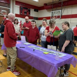 Group of students and adults stand around a long table with purple tablecloth at a school gym during a community fair or information night, looking at materials laid out on the table.