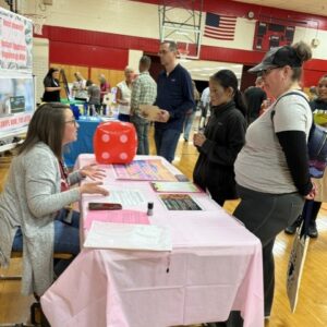 Volunteer at a pink-covered table in a gym answers questions from visitors; a large red die sits on the tabletop while people line up nearby.