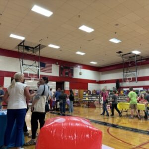 People at a community fair in a school gym; tables set up as booths along the court, with a red inflatable obstacle in the foreground.