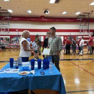 Two adults converse at a community event in a gym; a blue-clothed table with cups and papers is in the foreground.